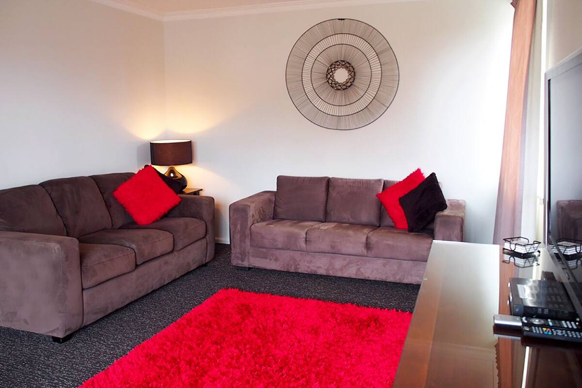 Un salon avec deux canapés, un tapis rouge, un ventilateur mural et un meuble pour la télévision.