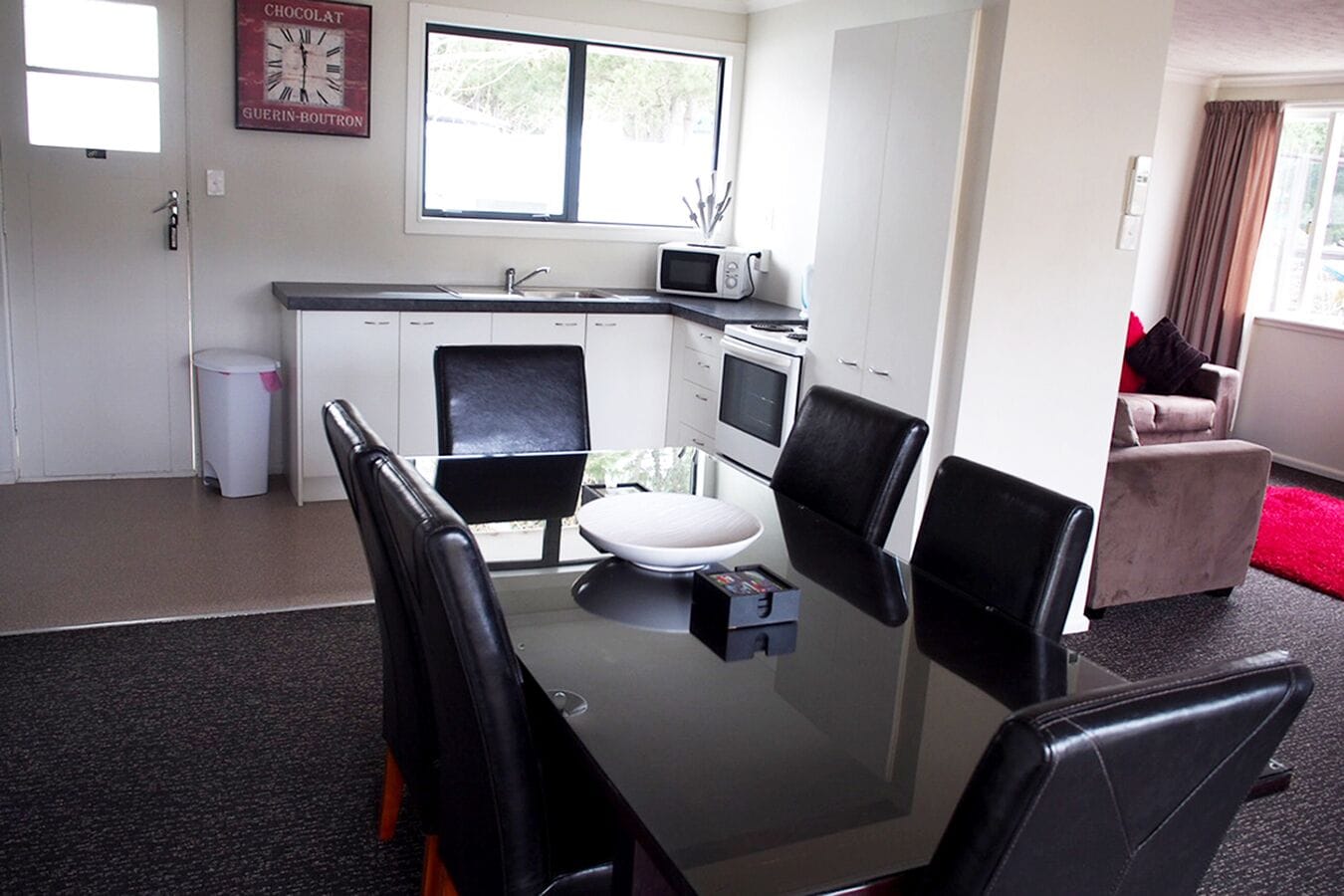 A modern kitchen with white cabinets, a microwave, and a sink. There is a dining table with four chairs and a white bowl. A trash can is visible near the door.