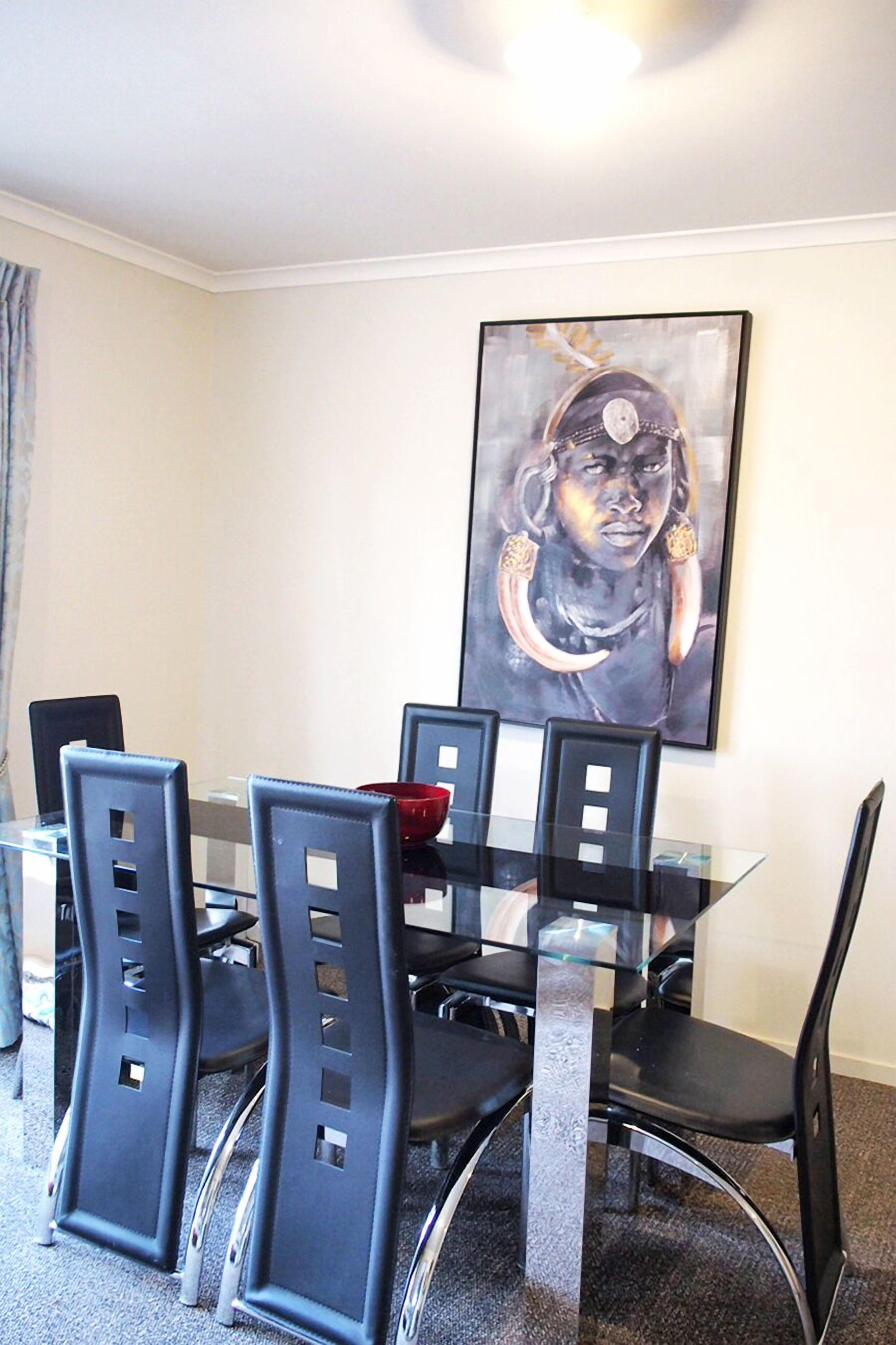 A dining area with a glass table and black chairs, a red bowl on the table, and a framed artwork on the wall.