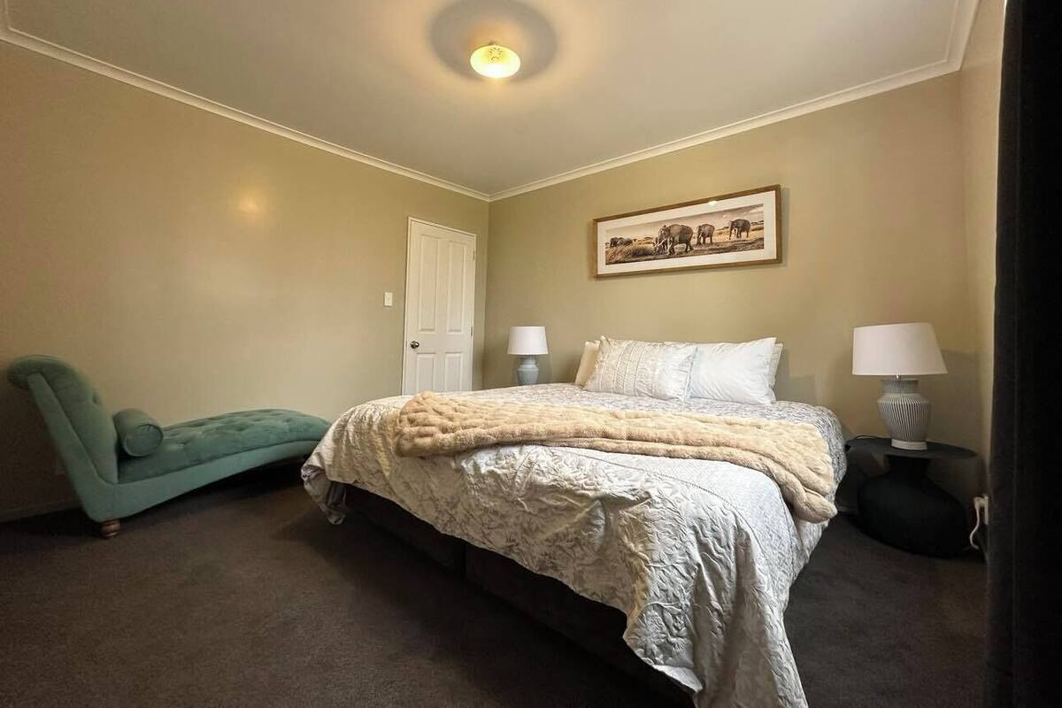 Une chambre à coucher avec un lit, deux lampes de chevet, une porte, un tableau encadré au mur et un fauteuil de couleur turquoise.