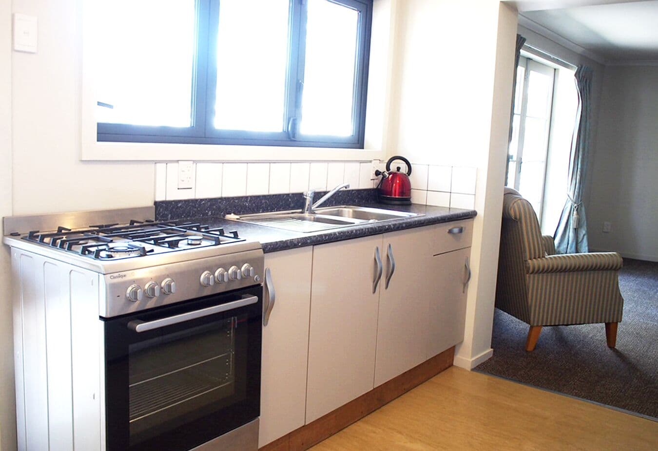 A compact kitchen with a gas stove, oven, and sink. There is a window above the sink and a chair in the adjacent living area.