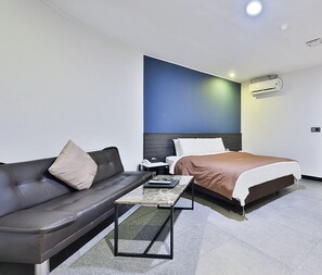 A modern hotel room with a dark leather sofa, a single bed with a brown bedspread, a small bedside table with a lamp, and a wall-mounted air conditioning unit. - Hotel Sopoong (Incheon)