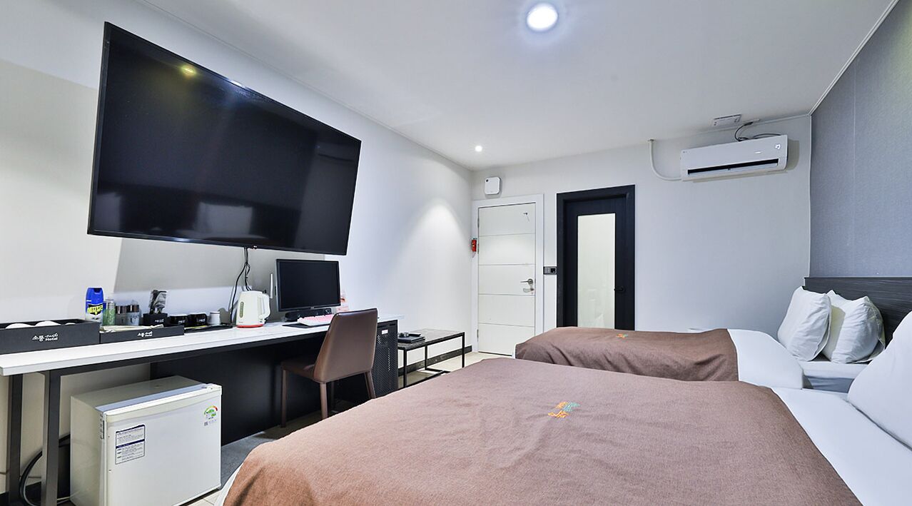 A hotel room with a bed, a desk with a computer, a TV, and an air conditioning unit.