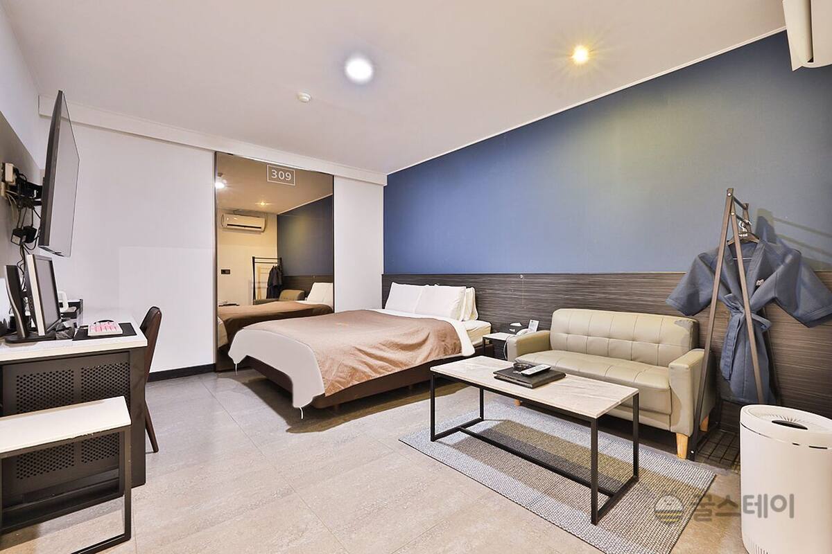 A modern hotel room with a bed, a sofa, a desk with a computer, and a television.