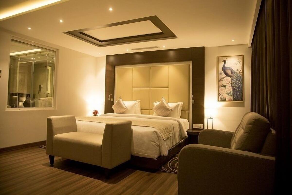 A modern hotel room with a large bed, two armchairs, a sofa, a large window, and a peacock painting.