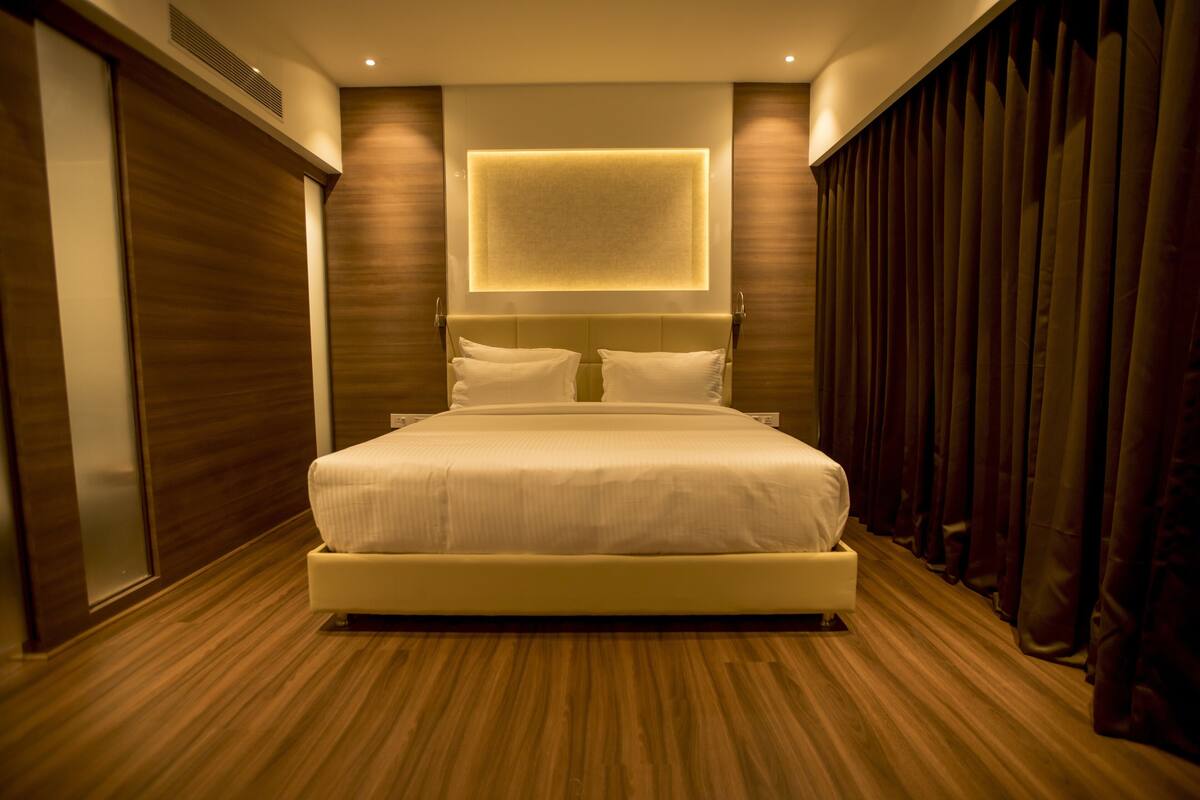 A modern hotel room with a large bed, wooden flooring, a wall-mounted light, and dark curtains.