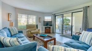 A living room with a sofa, armchair, and coffee table. There is a television and a sliding glass door leading to an outdoor area. - CRC 1112 - Steps Away from the Oceanfront Pool and Beach (St Augustine)