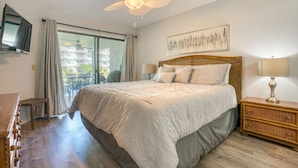 A bedroom with a large bed, a ceiling fan, a television, and a balcony with a view of buildings. - CRC 1112 - Steps Away from the Oceanfront Pool and Beach (St Augustine)