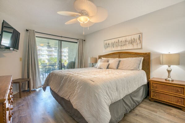 A bedroom with a large bed, a ceiling fan, a television, and a balcony with a view of buildings.