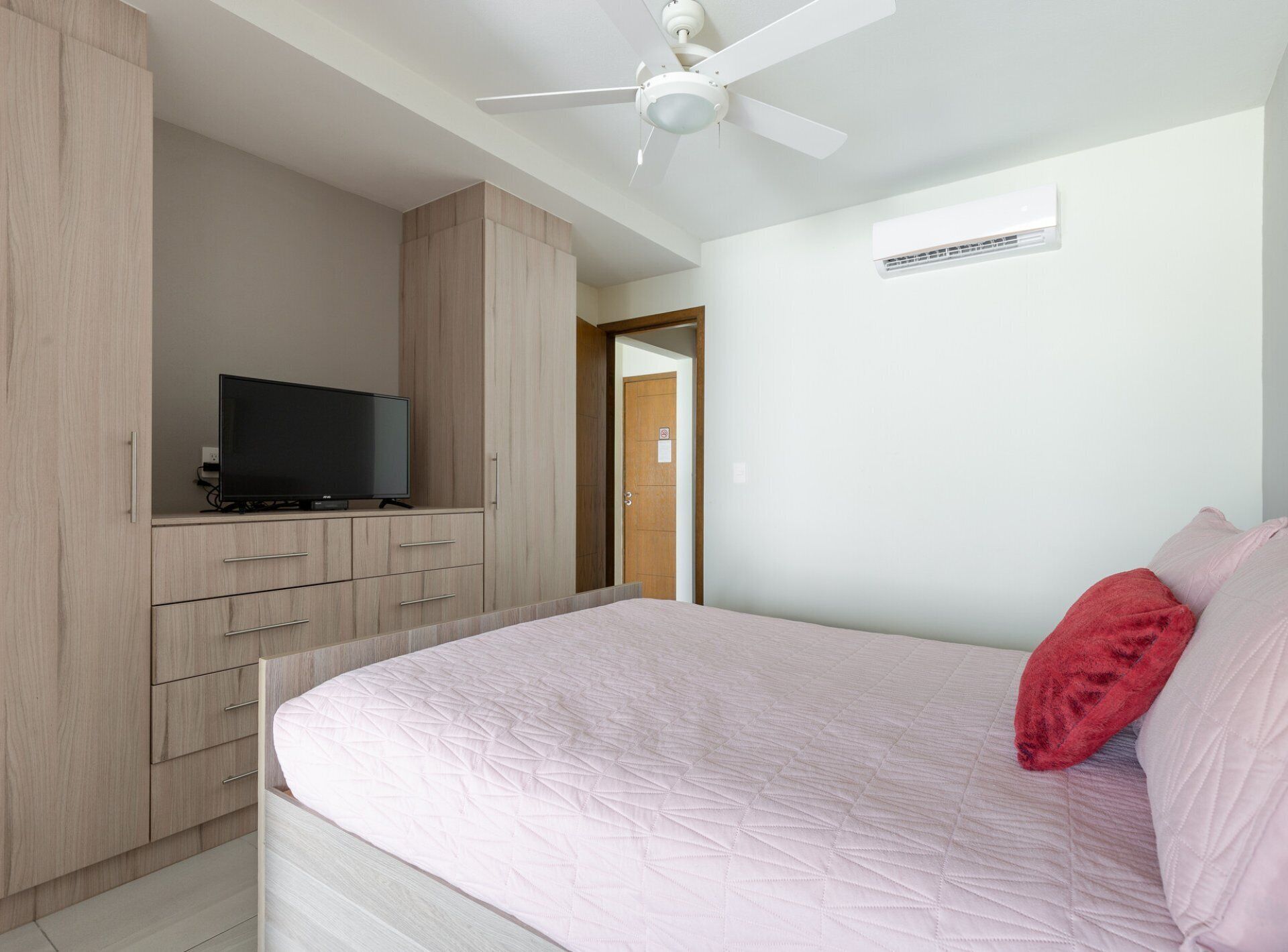 Ein Schlafzimmer mit einem Bett, einem Fernseher auf einer Holzkommode, einem Deckenventilator und einer Klimaanlage.