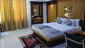 A hotel room with a bed, a desk, and a chair. There is a window with curtains, a wall-mounted air conditioner, and a decorative mirror.