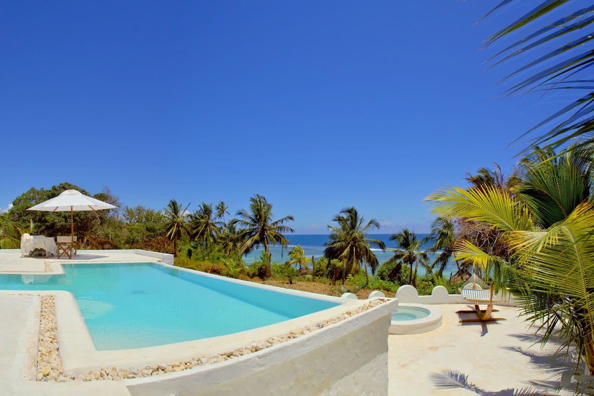 Zona de piscina con sillas de descanso y sombrillas, rodeada de palmeras y vista al océano.