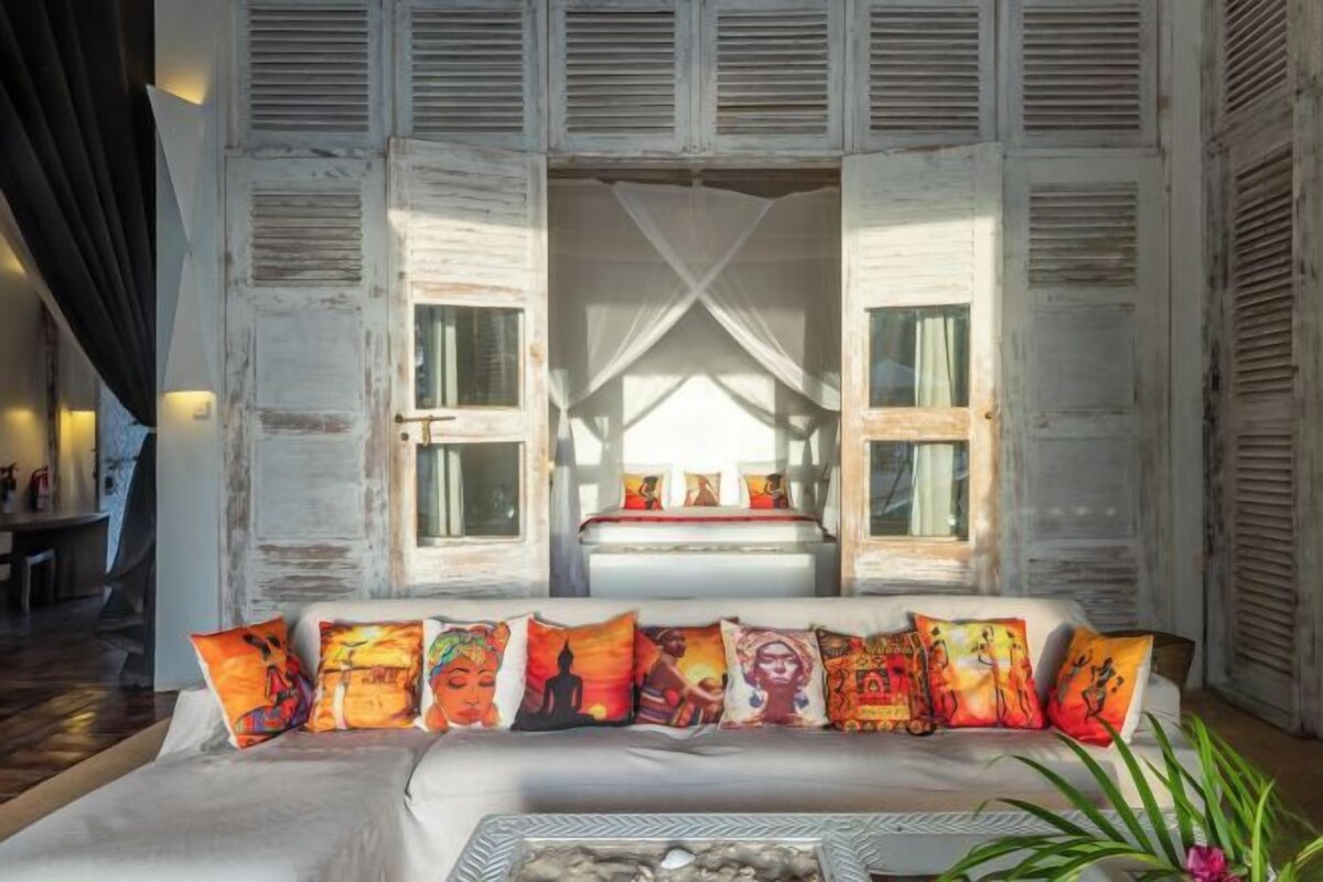 A cozy living room with a white sofa adorned with colorful cushions, a coffee table, and a view through a window with shutters.