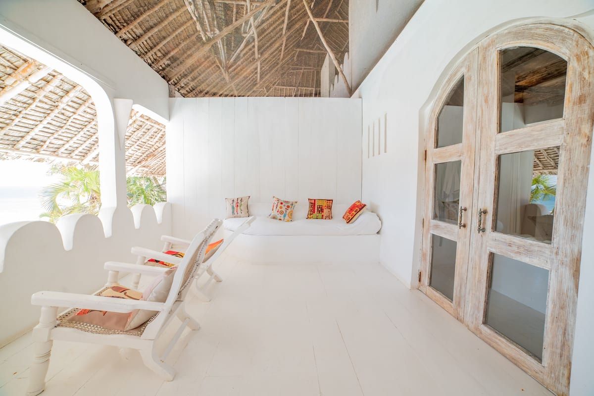 Una habitación con paredes blancas, techo de madera y un amplio ventanal arqueado. Hay bancos blancos con cojines de colores y un mueble de madera con puertas de cristal.
