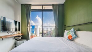 A hotel room with a large bed, a TV mounted on the wall, a small desk with a chair, and a balcony with a view of the city. - MH48 Hotel (Hengchun)