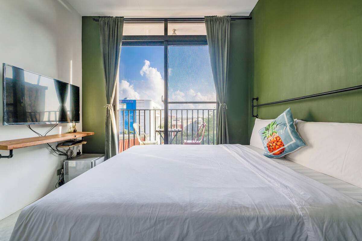 Habitació d'hotel amb un llit gran, una televisió penjada a la paret, un escriptori petit amb una cadira, i un balcó amb vista a la ciutat.