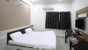 A hotel room with a bed, a desk with a chair, a television, an air conditioning unit, and a window with curtains. - Shree Hotel (Danta)