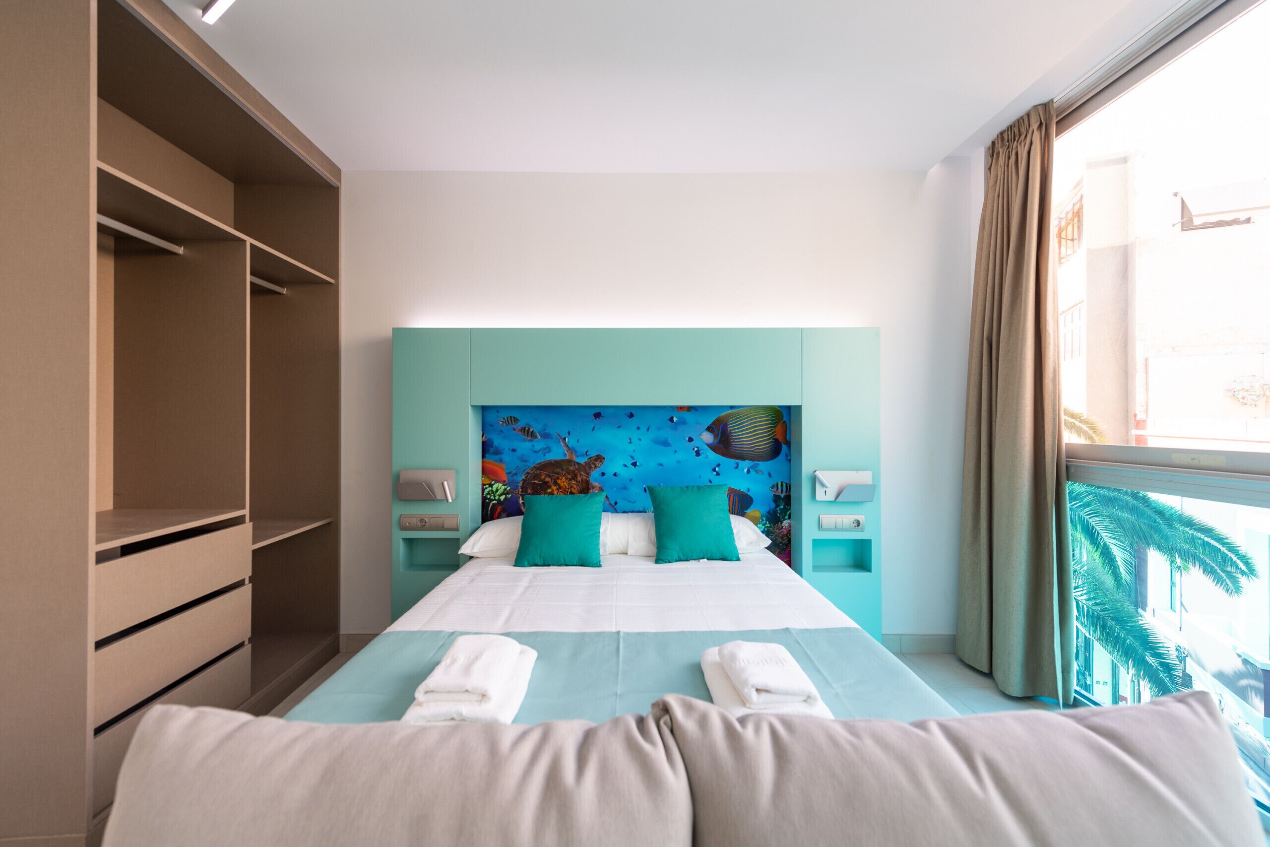 Una habitación de hotel con cama, cabecera con un mural, armario y ventana con cortinas.