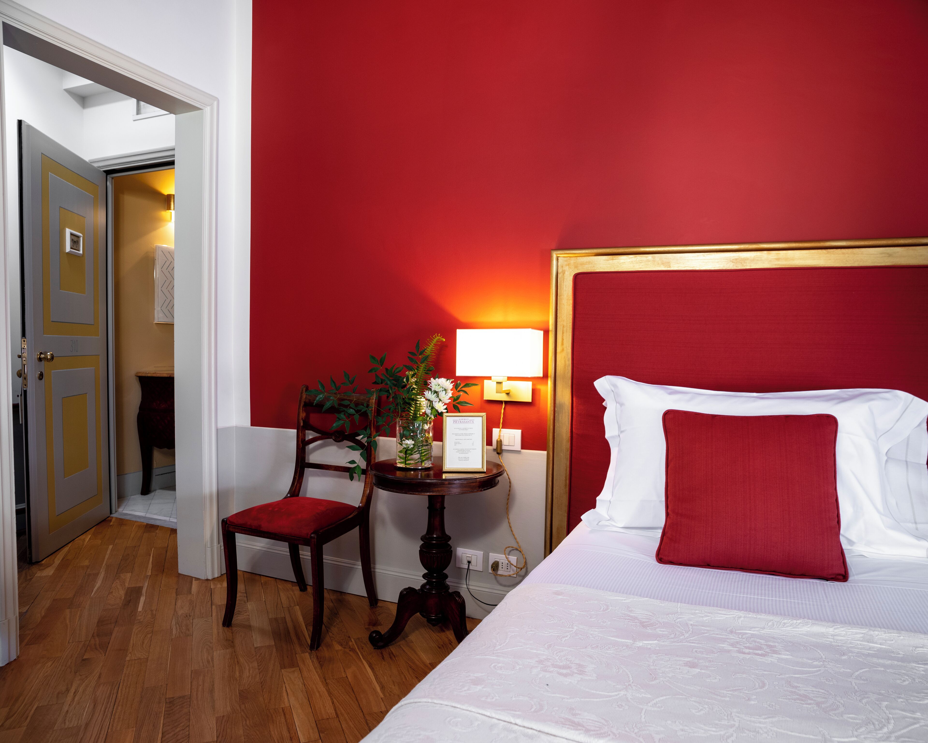 A hotel room with a red wall, a wooden chair with a red cushion, a bedside table with a lamp, and a bed with white bedding and a red pillow.