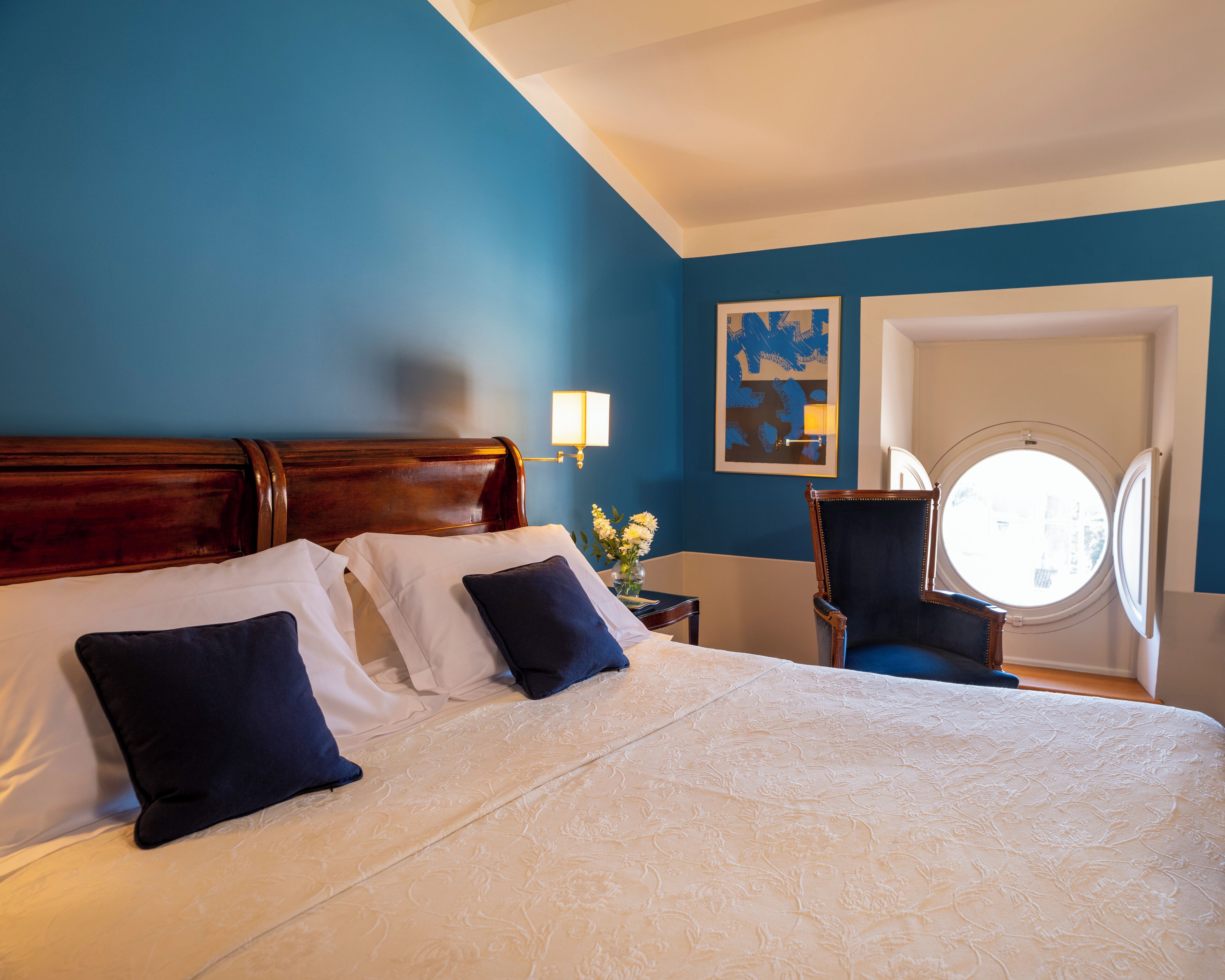 A hotel room with a large bed, a round window, and a blue wall.