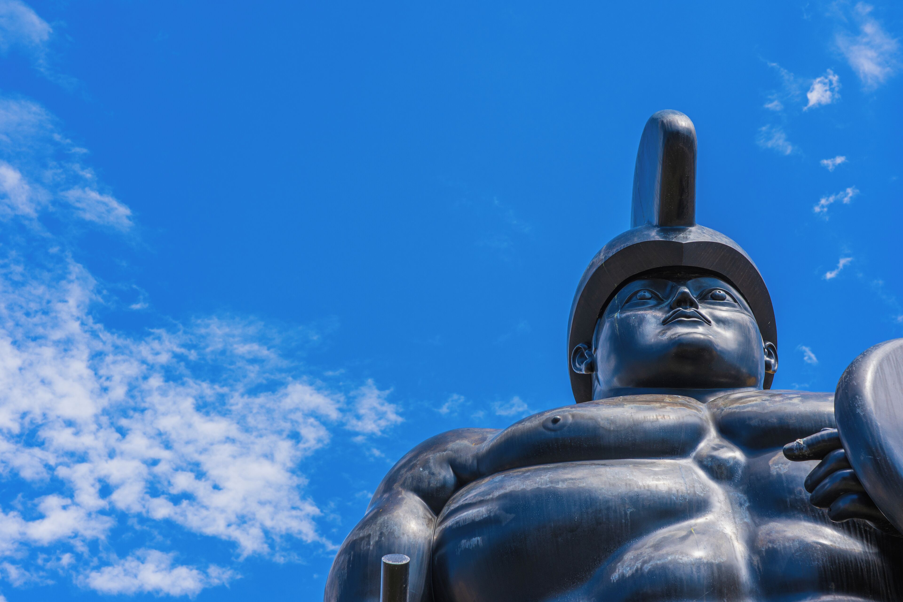 A large statue of a muscular figure with a helmet, set against a blue sky with clouds.
