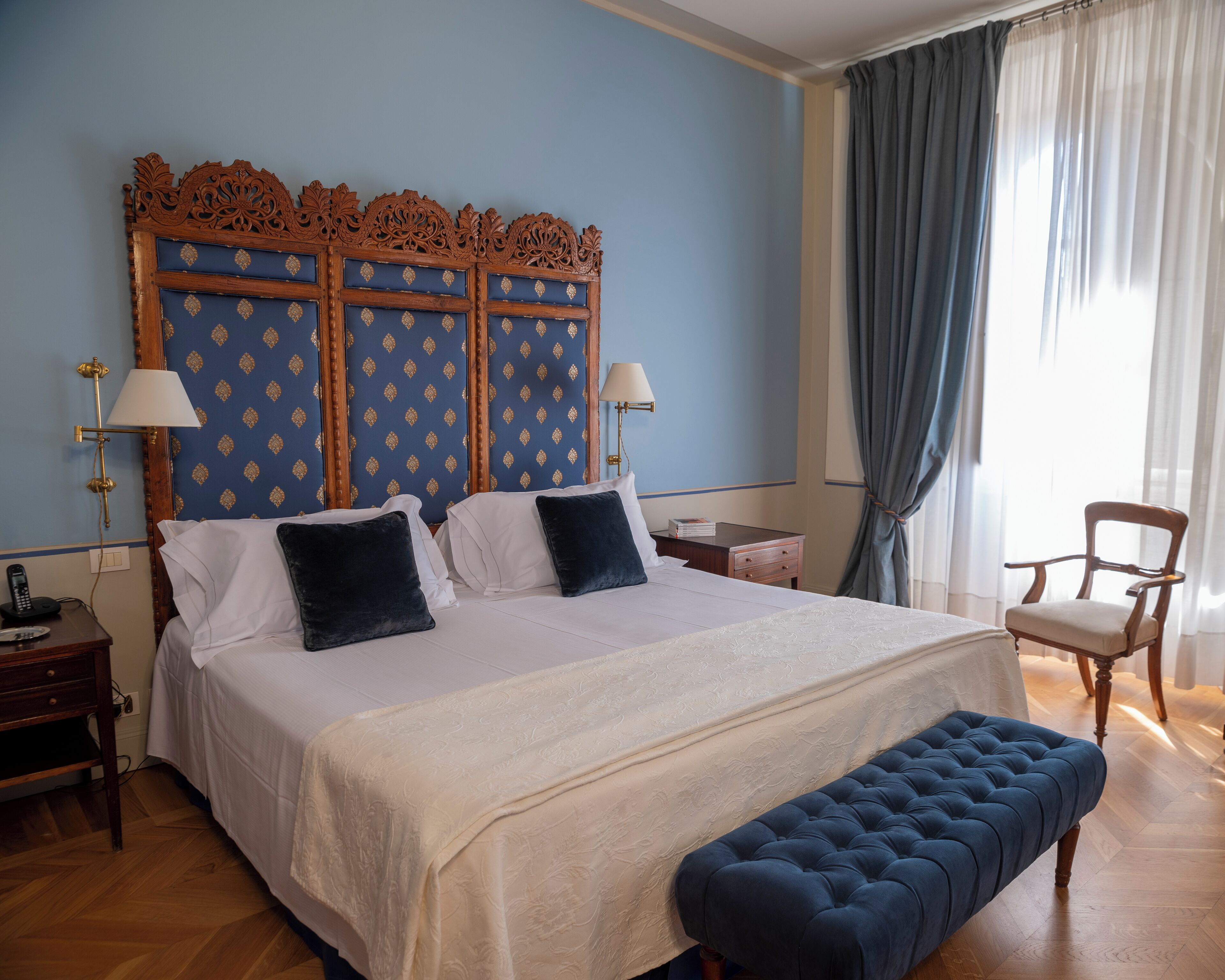 Une chambre d’hôtel avec un grand lit, une tête de lit en bois, deux tables de chevet avec des lampes, une chaise et une fenêtre avec des rideaux.