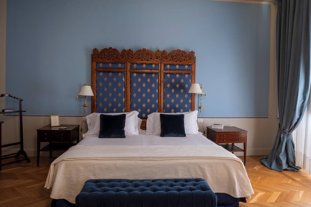 A bedroom with a large bed, two bedside tables, a nightstand, a blue upholstered bench, and a wooden headboard with intricate carvings.