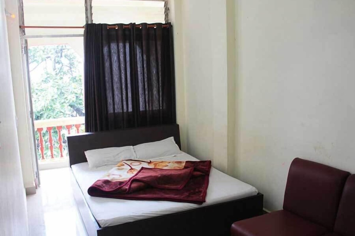 A single bed with a dark headboard, a white mattress, and a red blanket. There is a window with black curtains and a view of greenery outside.