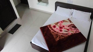A bed with a floral bedspread, white pillows, and a wooden headboard in a room with white walls and a tiled floor. - Hotel Banjara (Pipariya)