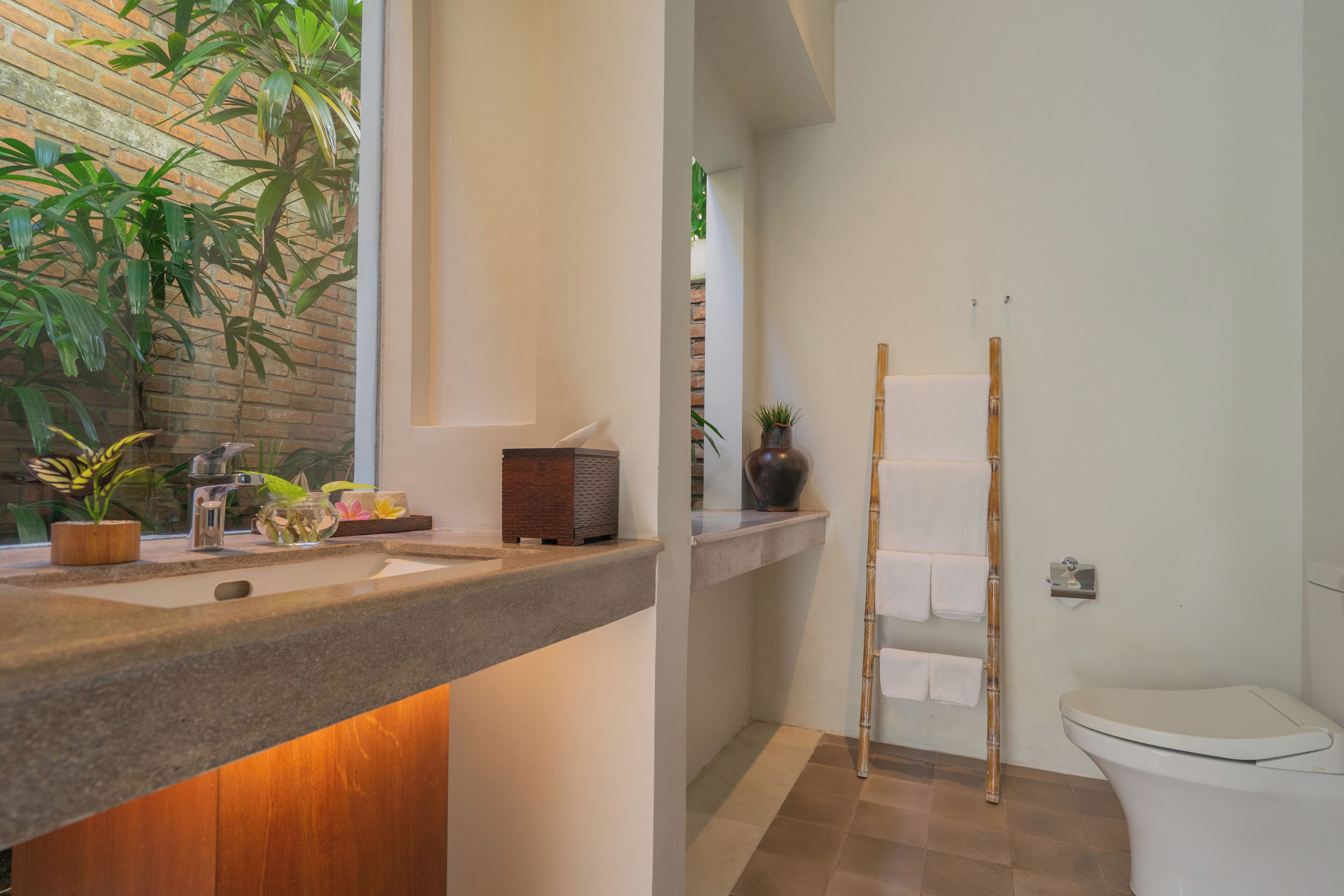 Un baño con lavamanos, espejo, toallero de bambú, inodoro y ventana con plantas.