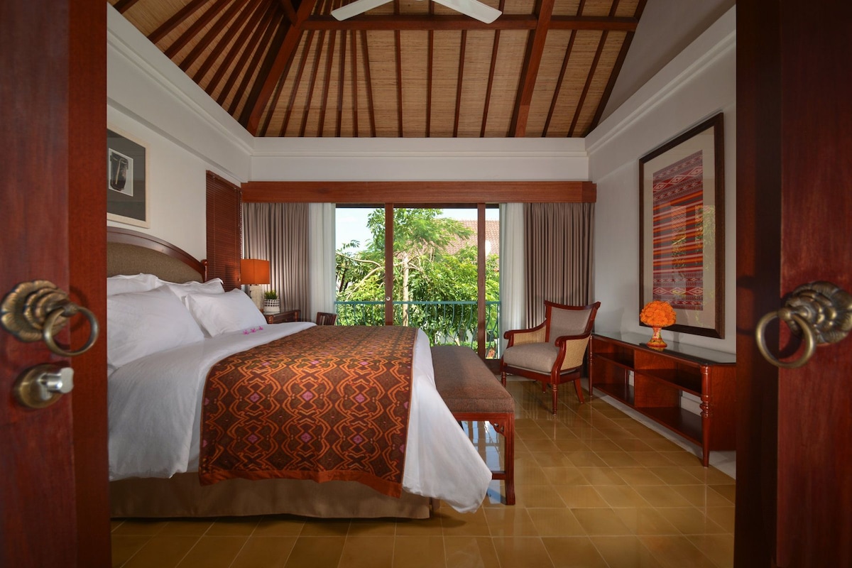 A hotel room with a large bed, a desk with a chair, a balcony with a view of greenery, and a ceiling fan.