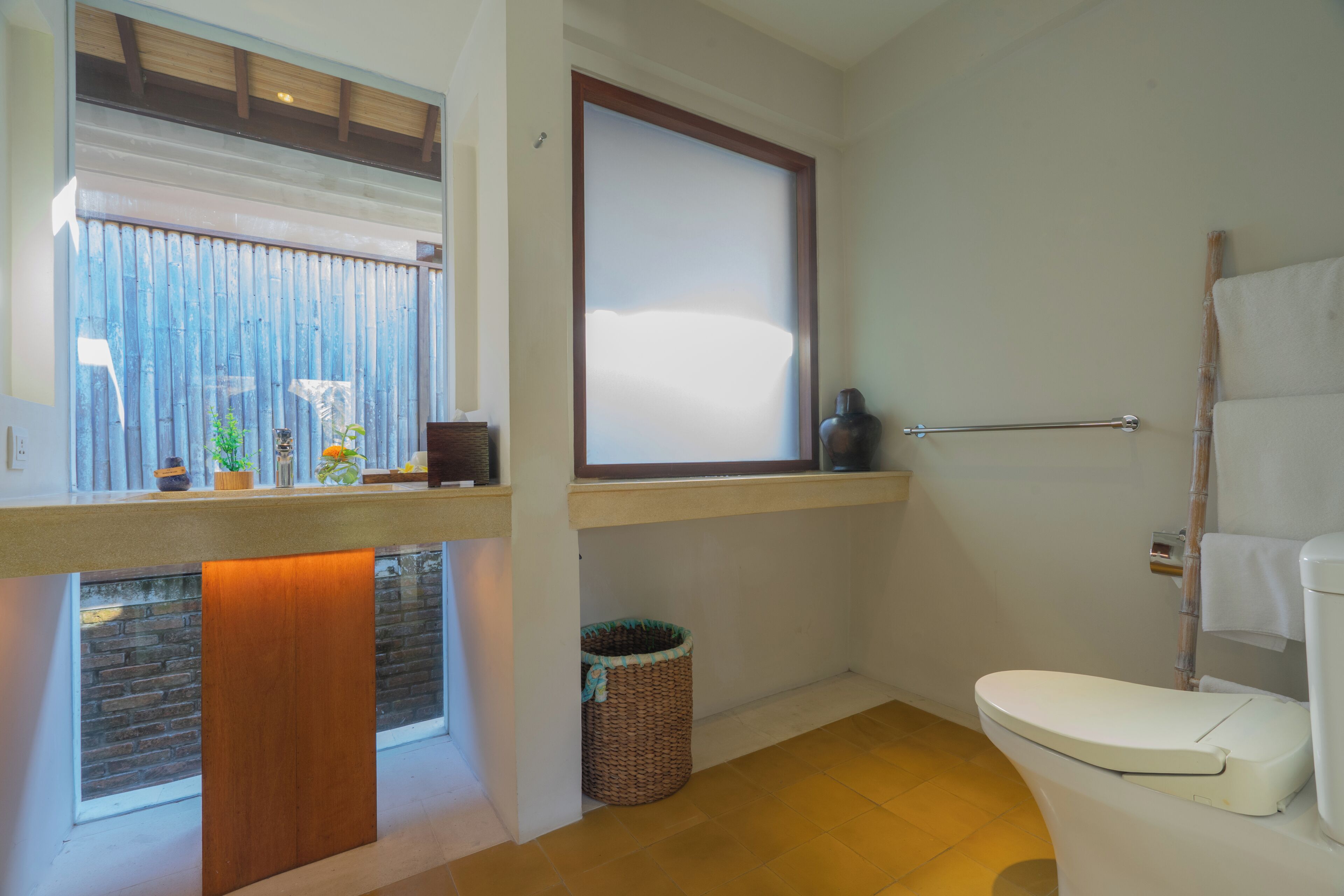 A bathroom with a toilet, a window with blinds, a wicker basket, and a shelf with small items.