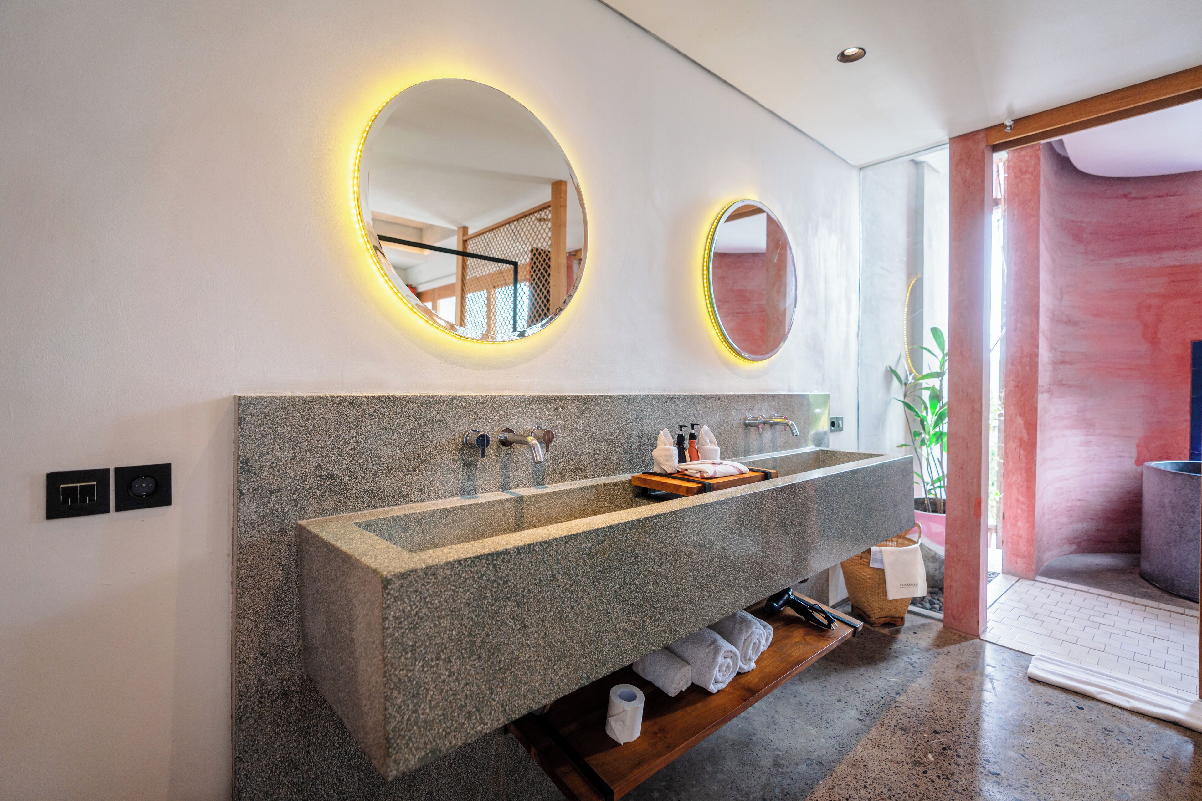 Un baño moderno con un lavabo de granito, dos espejos con iluminación y un estante de madera con toallas.