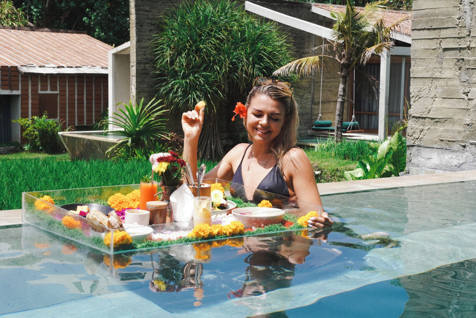 A person sitting by a glass table with a tray of food and drinks, surrounded by greenery and a building in the background.