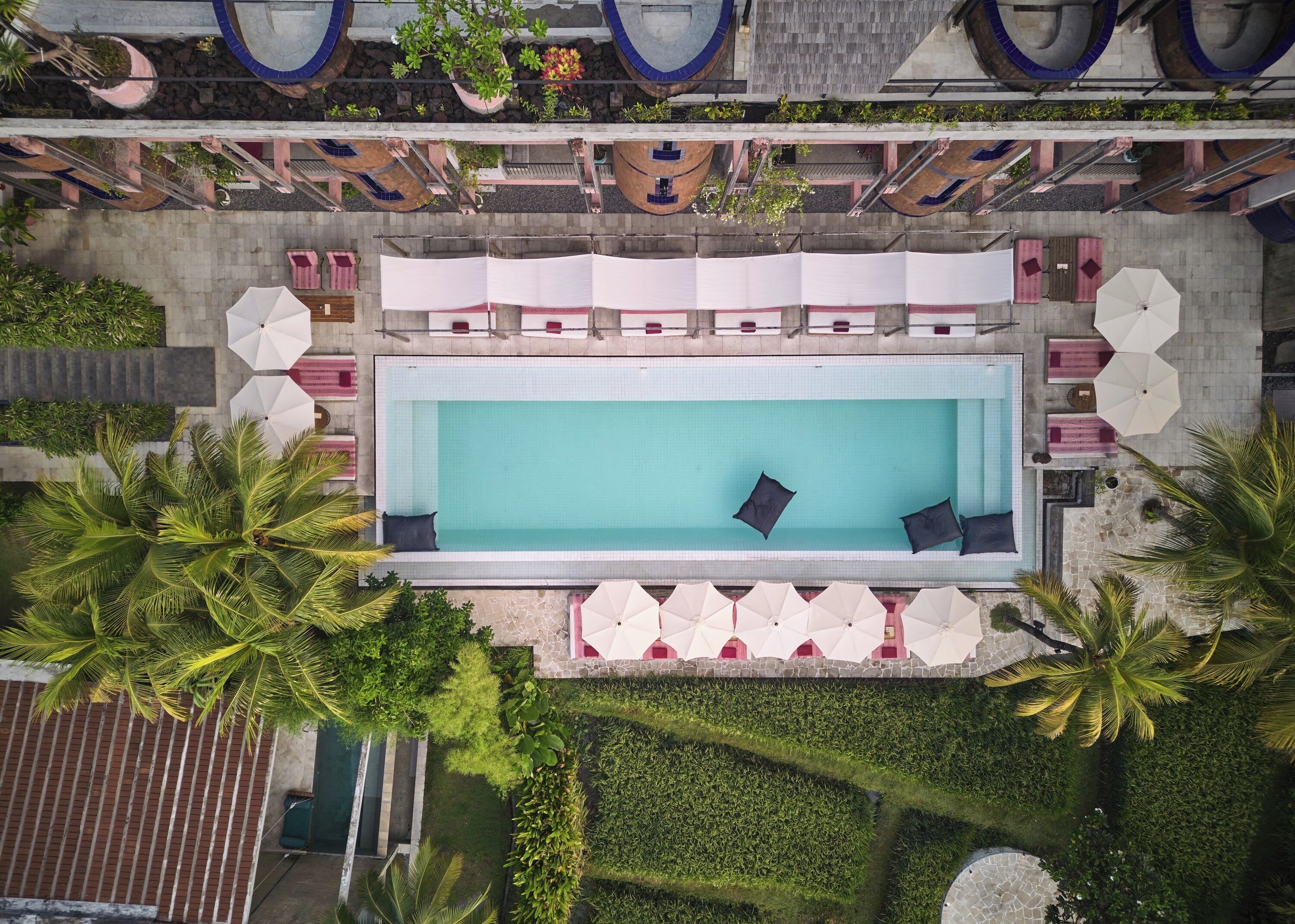 An aerial view of a hotel with a rectangular pool surrounded by lounge chairs and umbrellas, palm trees, and a stone facade.