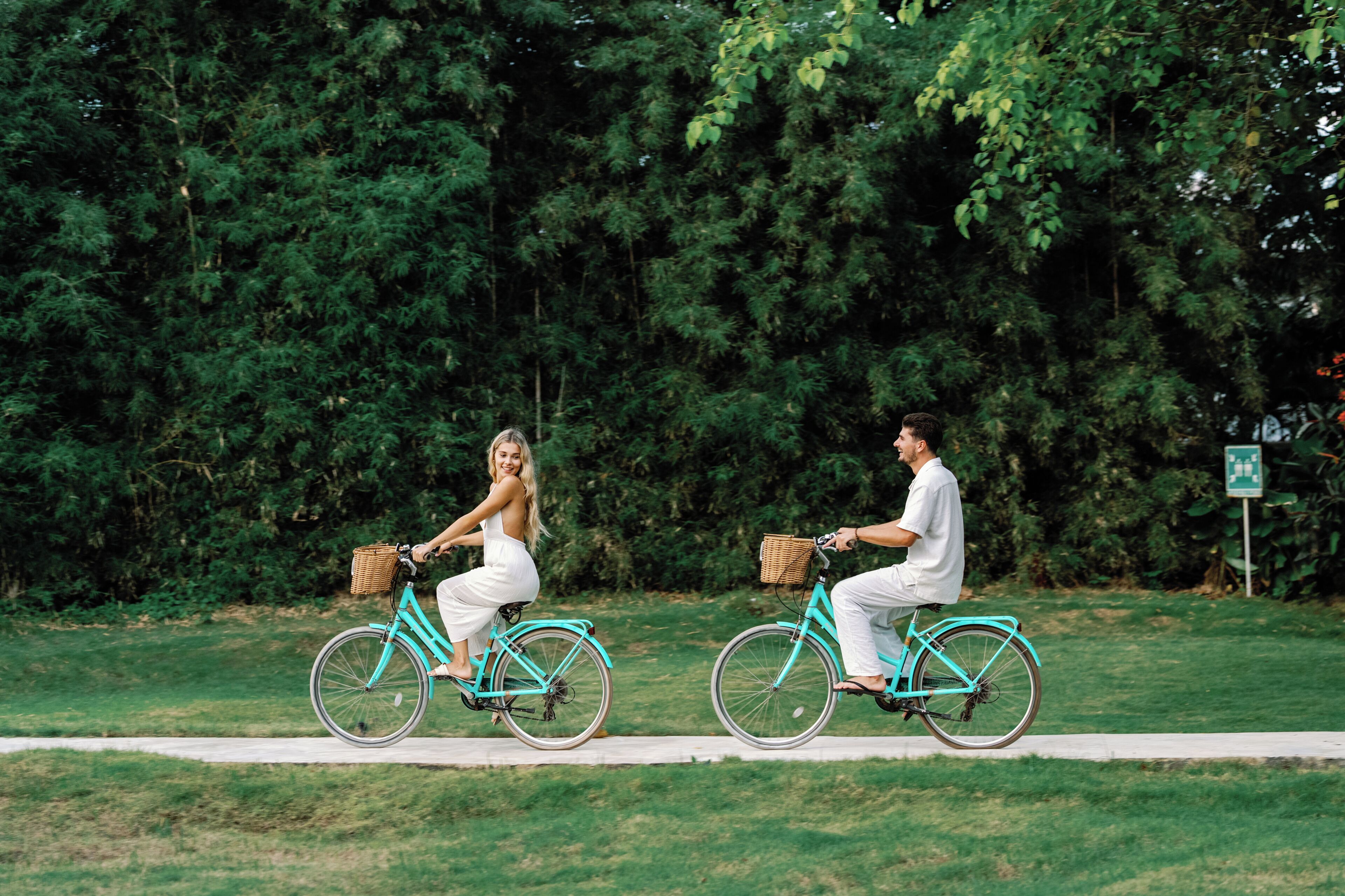 Two people riding bicycles on a path with a dense green hedge in the background.