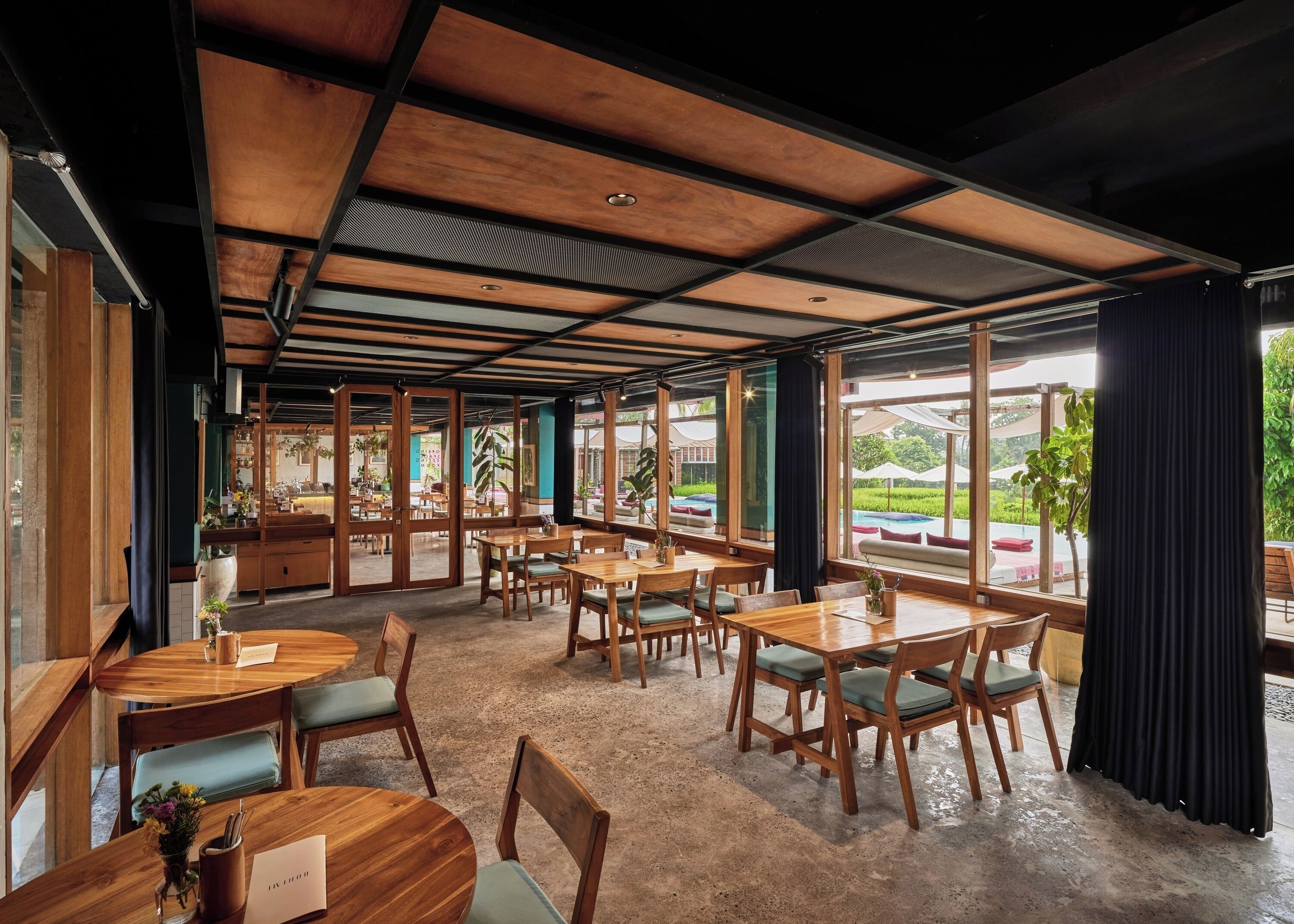 A spacious indoor area with wooden tables and chairs, large windows offering views of a garden and pool, and a high ceiling with exposed beams.
