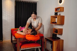 A massage therapy session in a room with a tiled wall, a black curtain, and wooden shelves with various items.