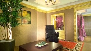 Habitación de hotel con una planta grande en maceta, una mesa de comedor con alfombra roja, una silla negra y un cuadro en la pared.