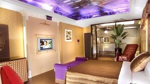 Una habitación de hotel con una cama grande, televisión, una silla roja y un banco púrpura.