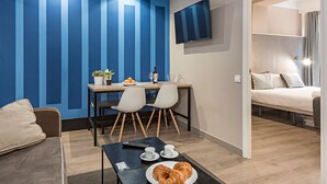 A modern hotel room with a blue accent wall, a flat-screen TV, a dining area with a table and chairs, a bed, and a coffee table with croissants. - Cool Jazz 24 - Habitat Apartments (Barcelona)