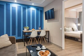 A modern hotel room with a dining area, a flat-screen TV, a bed, and a breakfast setup with croissants. - Cool Jazz 24 - Habitat Apartments (Barcelona)
