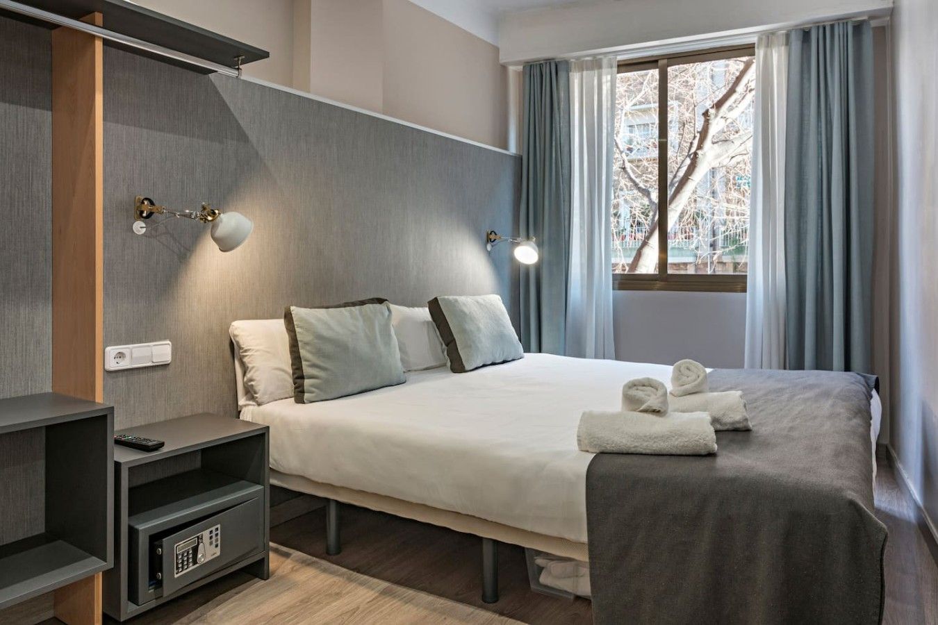 Ein modernes Hotelzimmer mit Bett, Nachttisch und Blick auf Bäume durch das Fenster.