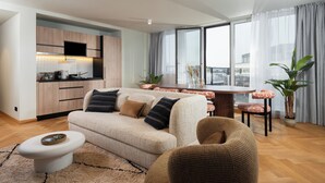 A modern living room with a sofa, coffee table, and dining area. There is a kitchen with wooden cabinets and a flat-screen TV. The room has large windows with curtains and a view of the city. - master Mirabell (Salzburg)
