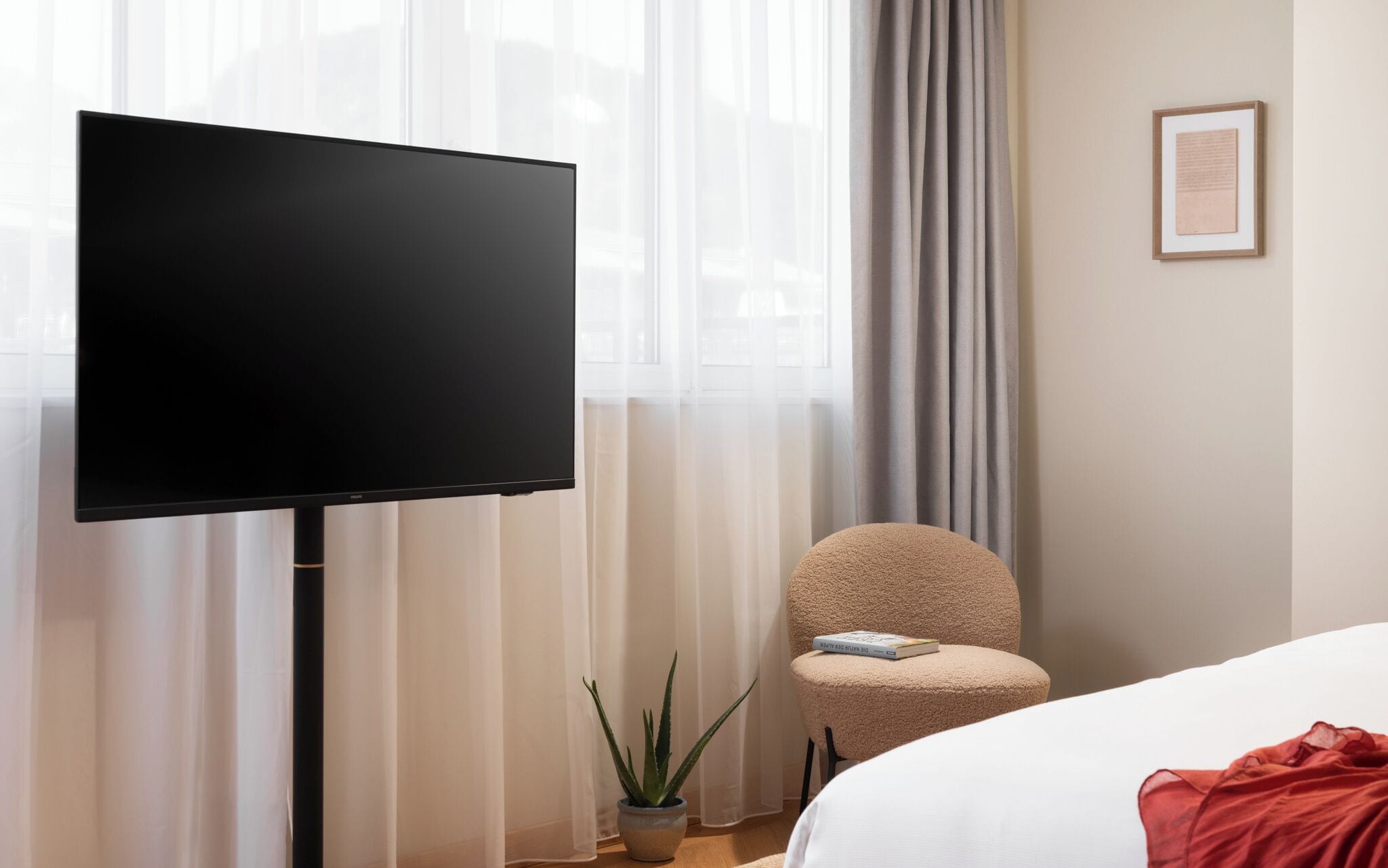 A modern hotel room with a large flat-screen TV mounted on the wall, a comfortable armchair, a small potted plant, and a neatly made bed.