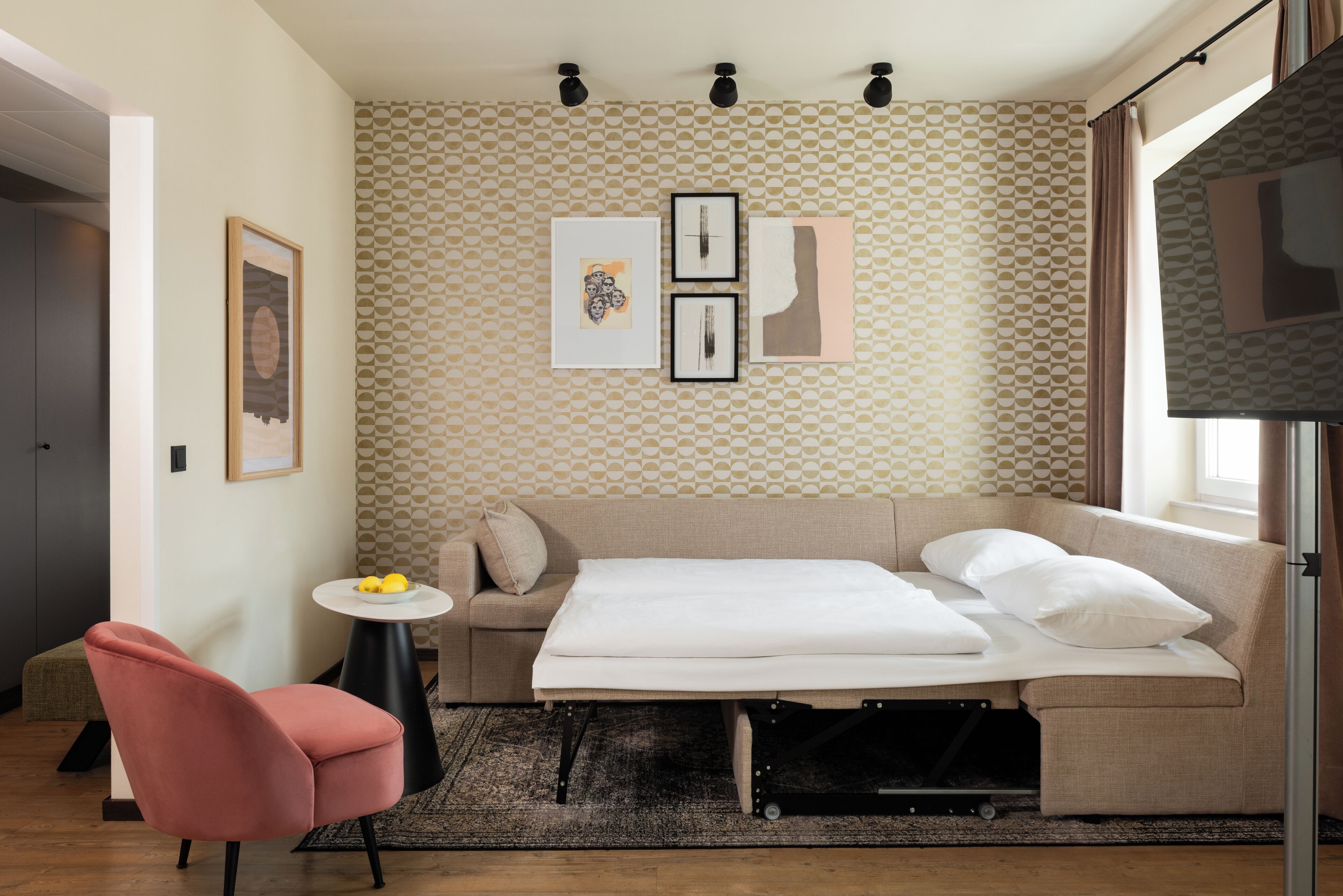 Een moderne hotelkamer met een slaapbank, een klein rond tafeltje met een schaal fruit, een roze stoel en een televisie rechts.