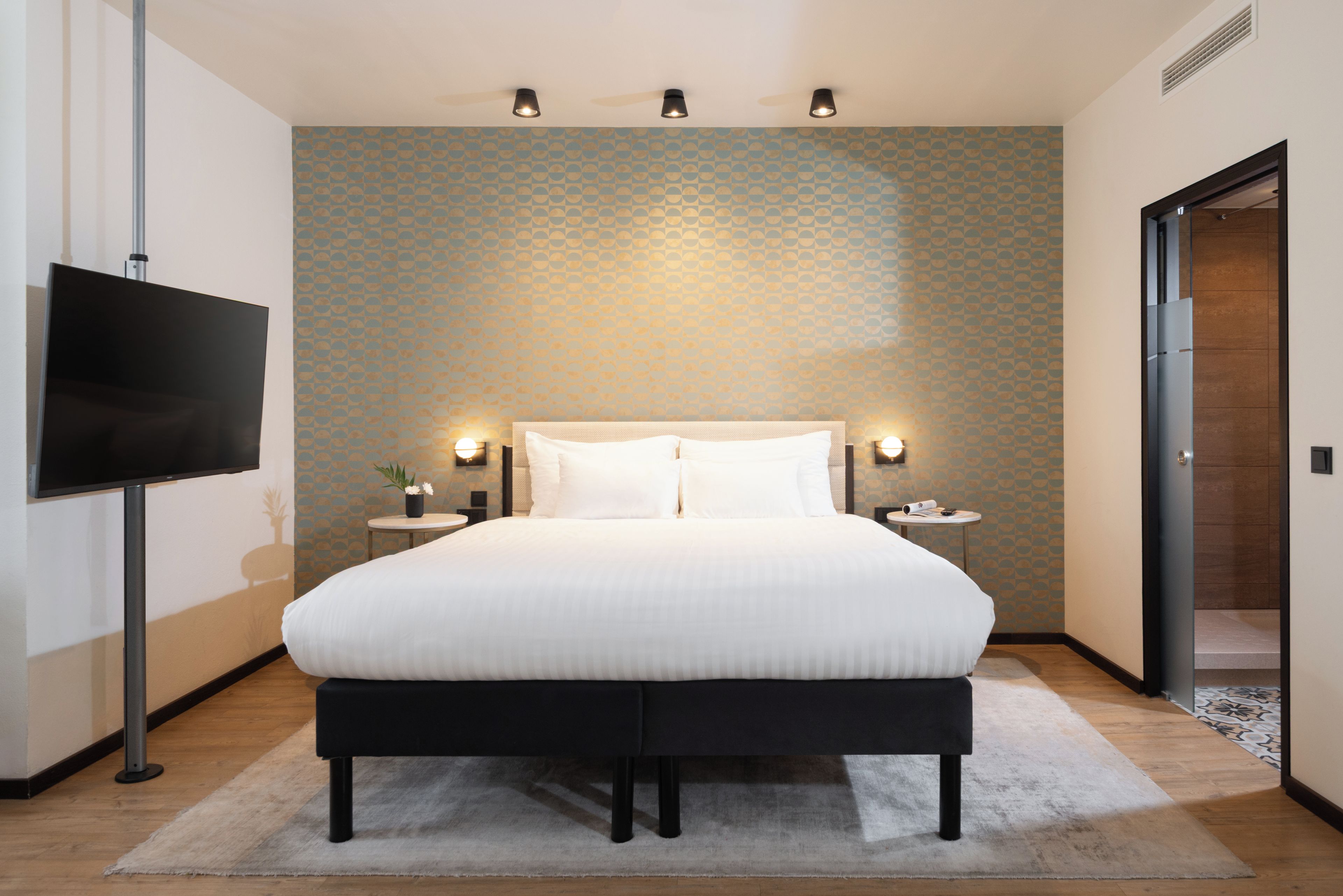 A modern hotel room with a large bed, a flat-screen TV, bedside lamps, and a patterned wall.