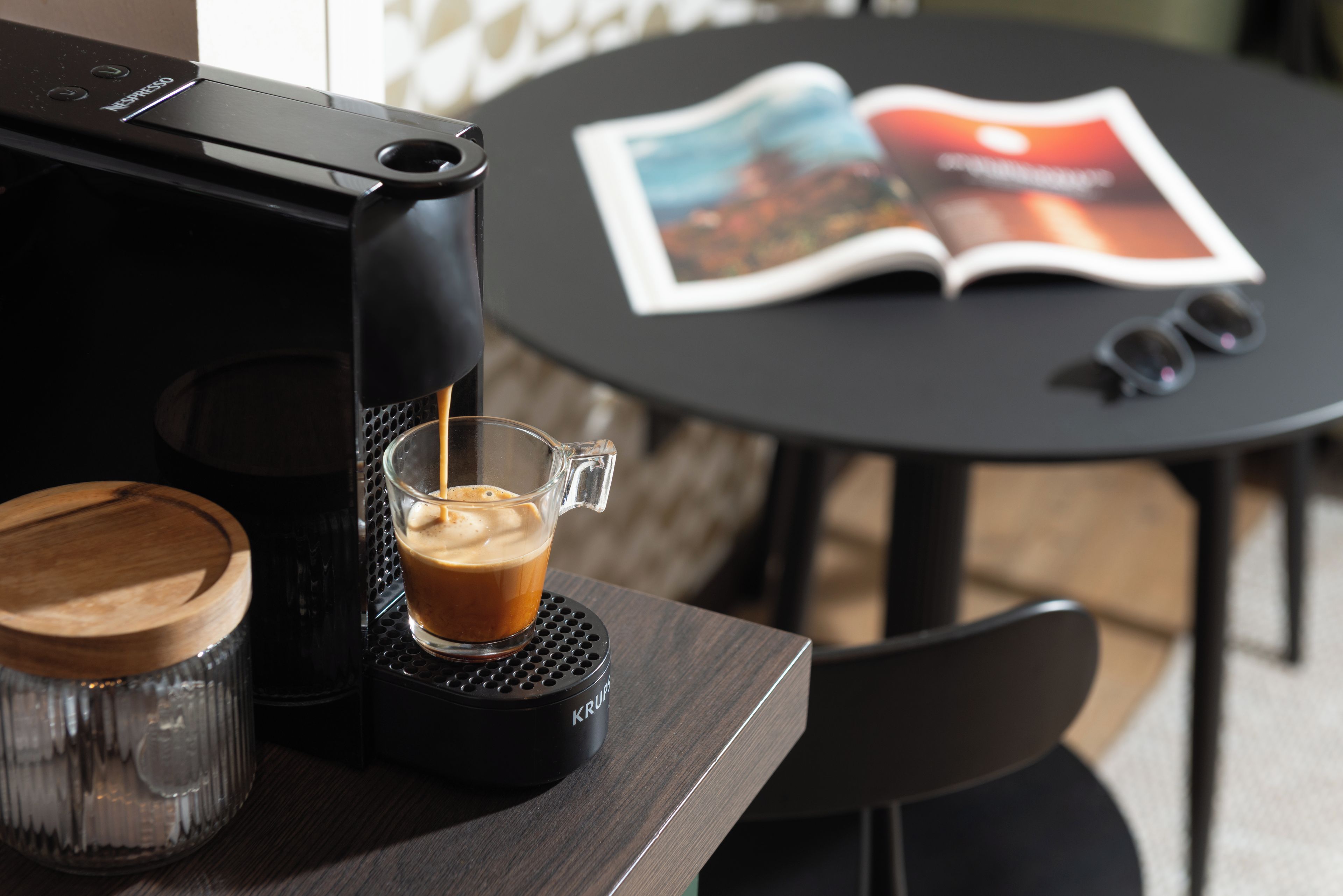 A coffee machine dispensing coffee into a glass cup on a coaster, with a magazine on a table in the background.