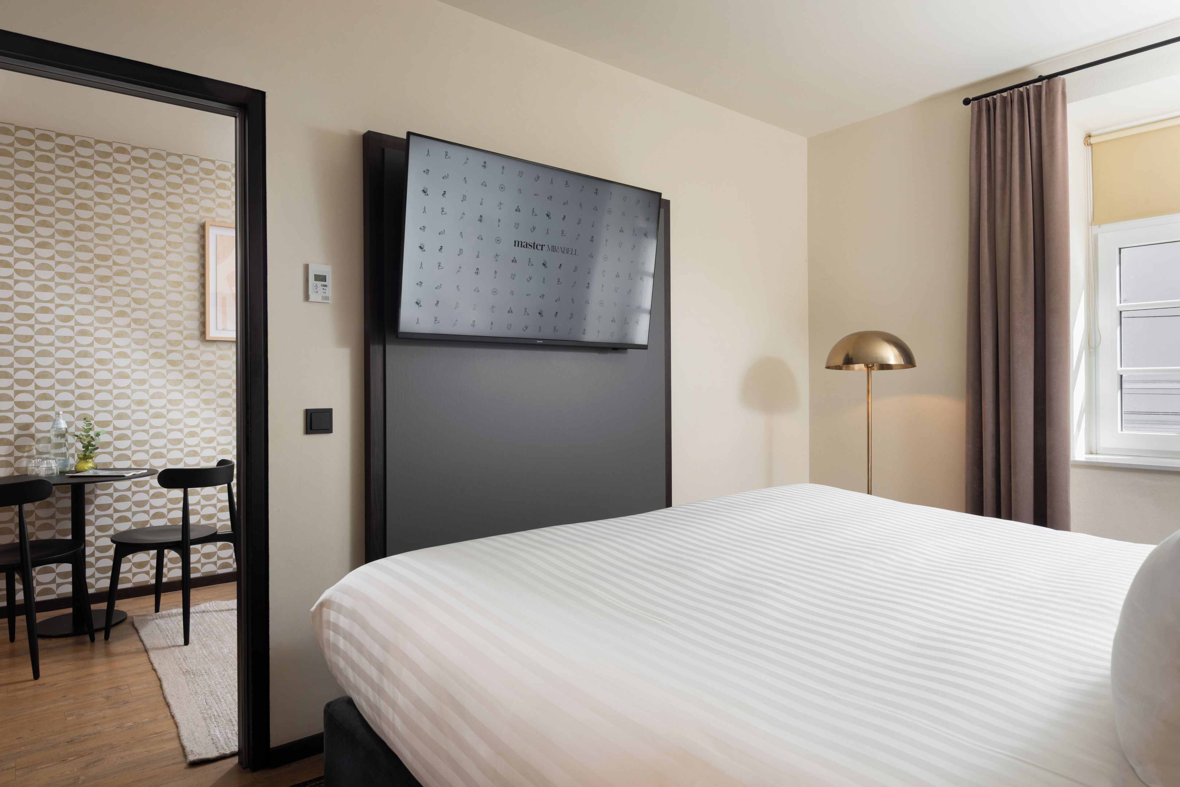Ein Hotelzimmer mit einem Bett, einem Schreibtisch mit Stühlen, einer Stehlampe und einer wandmontierten Bedieneinheit.