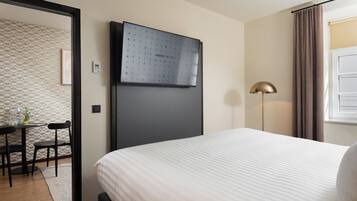 Ein Hotelzimmer mit einem Bett, einem Schreibtisch mit Stühlen, einer Stehlampe und einer wandmontierten Bedieneinheit.